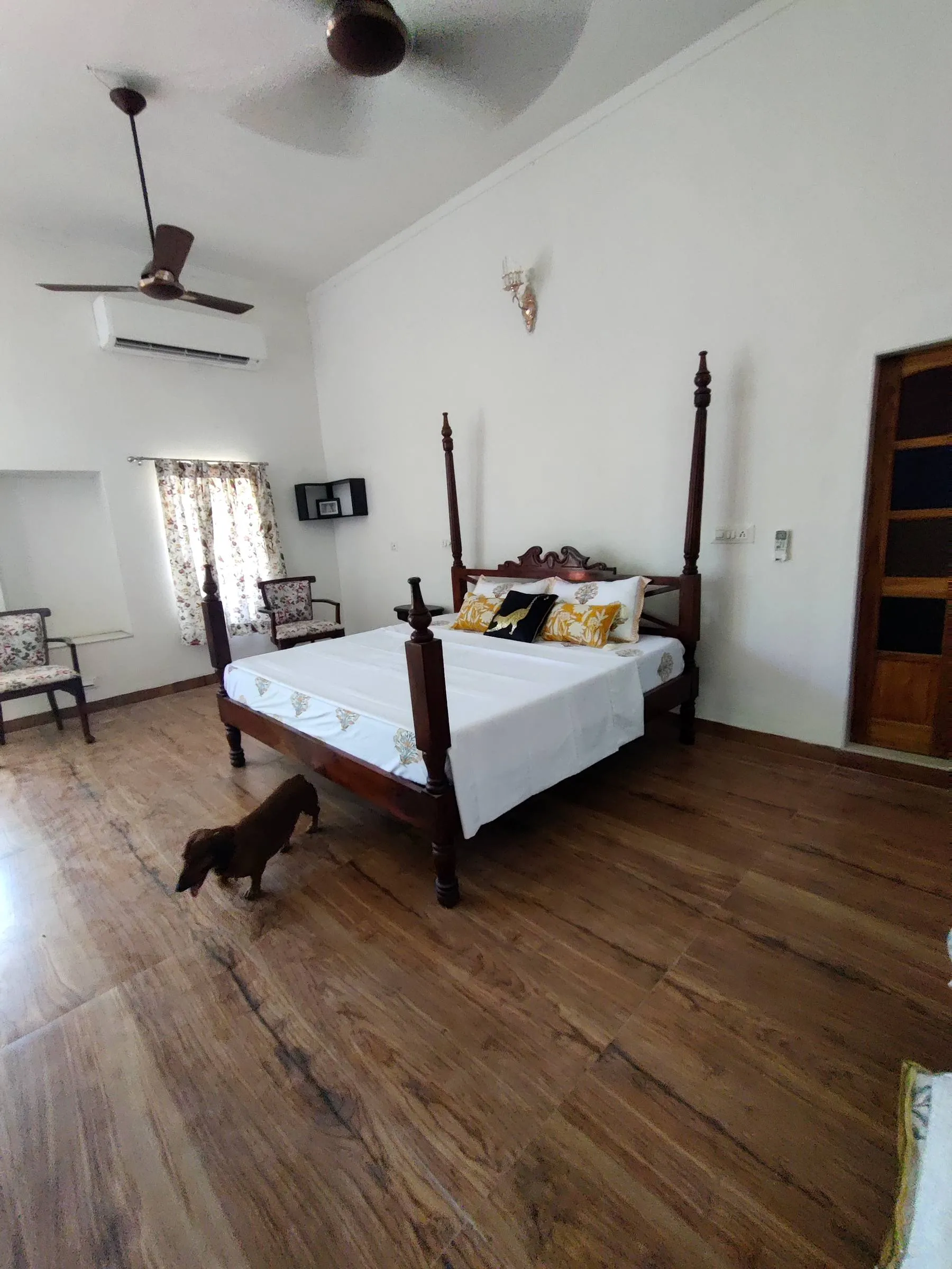 A tall, airy guest room at Rawla Bankli with a carved wooden four-poster bed on a wide-plank floor, a ceiling fan turning above